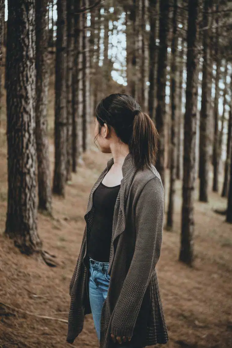 brown haired woman in jeans and woolen cardigan standing in a forest